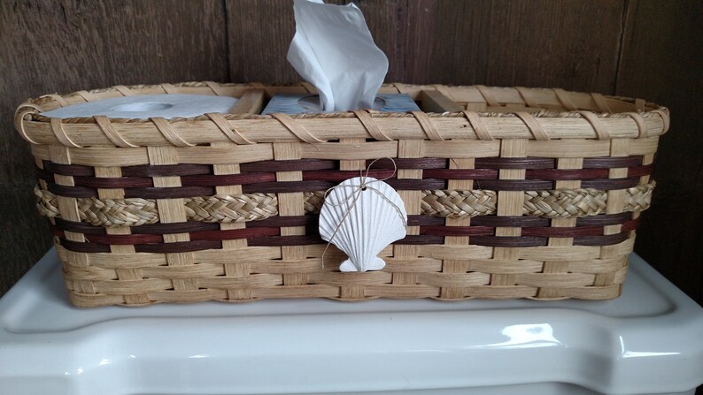 May include: A woven wicker basket with a brown and tan pattern. The basket has three compartments, one for a roll of toilet paper, one for a box of tissues, and one for other items. A white seashell is attached to the front of the basket.