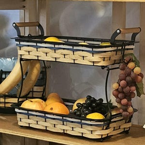 May include: Two-tiered fruit basket with a woven design and black metal frame. The basket has wooden handles and is filled with various fruits, including lemons, oranges, and grapes. The basket is designed to hold and display fruit.