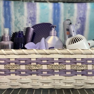 May include: A white wicker basket with purple accents, filled with hair care products, including a hair dryer, brushes, and bottles of hairspray.