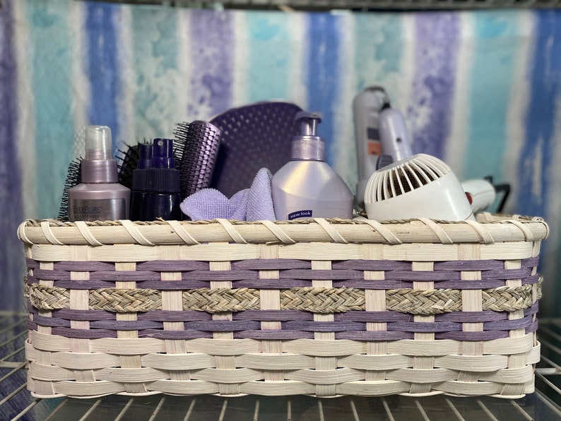 May include: A white wicker basket with purple accents, filled with hair care products, including a hair dryer, brushes, and bottles of hairspray.