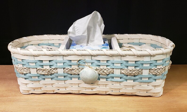 May include: White wicker tissue box holder with a blue and tan woven design. The holder has a seashell accent and is divided into three sections.