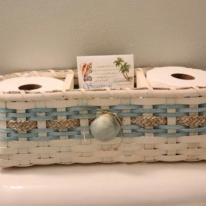 May include: White wicker bathroom tissue holder with two rolls of toilet paper and a decorative seashell. The holder has a blue and tan woven design and a card with a seashell and palm tree design.