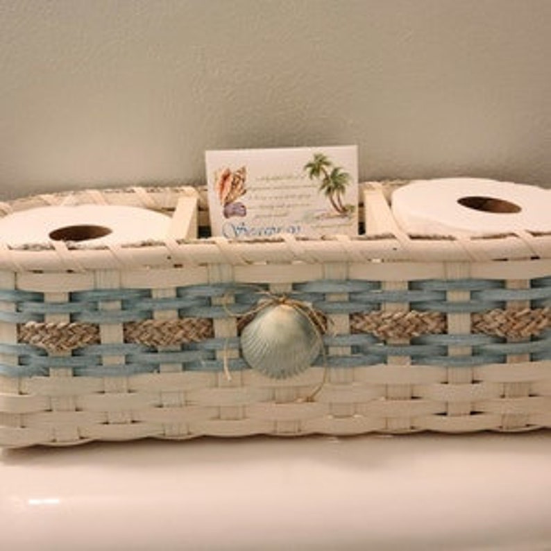 May include: White wicker bathroom tissue holder with two rolls of toilet paper and a decorative seashell. The holder has a blue and tan woven design and a card with a seashell and palm tree design.