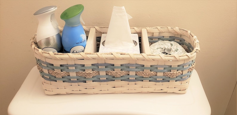 May include: A white wicker basket with blue and tan accents holds two spray bottles, a box of tissues, and a roll of toilet paper.