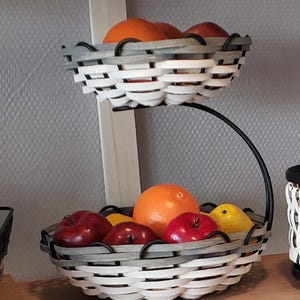 May include: A two-tiered fruit basket made of woven white and gray wicker with a black metal stand. The top tier holds oranges and the bottom tier holds apples and a yellow pear.