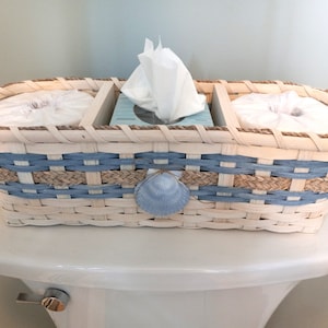 May include: A woven basket with blue and tan stripes holds three tissue boxes. The basket has a seashell design and is sitting on a white toilet tank.