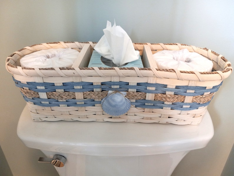 May include: A woven basket with blue and tan stripes holds three tissue boxes. The basket has a seashell design and is sitting on a white toilet tank.
