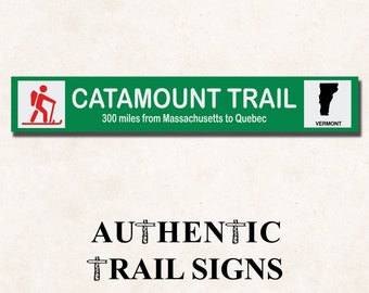 May include: Green trail sign with white text that reads "Catamount Trail 300 miles from Massachusetts to Quebec". A red stick figure hiker with a backpack and hiking stick is on the left side of the sign. A black silhouette of the state of Vermont is on the right side of the sign with the word "Vermont" below it. The text "Authentic Trail Signs" is below the sign.