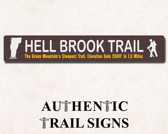 May include: A rectangular brown sign with white text reading HELL BROOK TRAIL, a white Vermont state outline, and a white hiker icon. Below, yellow text reads The Green Mountain's Steepest Trail, Elevation Gain 2600' in 1.5 Miles.
