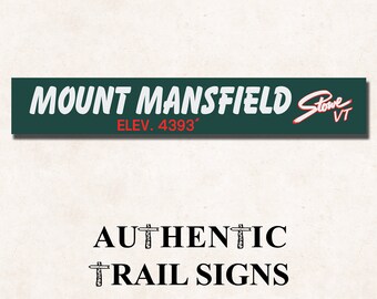 May include: A green and white sign with the text "MOUNT MANSFIELD Stowe VT ELEV. 4393'" in white lettering. Below the sign is the text "AUTHENTIC TRAIL SIGNS" in black lettering.