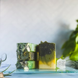 May include: Three bars of handmade soap, stacked on top of each other. The soap is green and yellow with a marbled pattern. The soap is sitting on a glass surface. The text "Hunter Homes" is visible on a clear crystal.