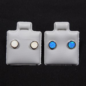Peut inclure: Deux paires de boucles d'oreilles clous sur une carte d'exposition grise. Une paire est en opale blanche et l'autre en opale bleue.