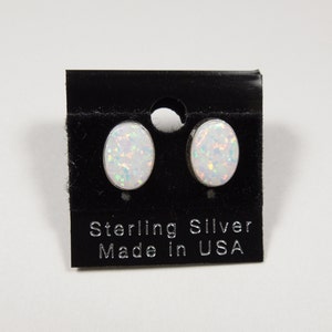 Könnte beinhalten: Ovale Opal-Ohrstecker aus Sterlingsilber, präsentiert auf einer schwarzen Karte. Auf der Karte steht "Sterling Silver Made in USA". Die Opale sind weiß mit schillernden Flecken, die einen Schimmereffekt erzeugen.