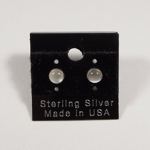 May include: A pair of silver stud earrings with white moonstone stones. The earrings are on a black card with the text "Sterling Silver Made in USA".