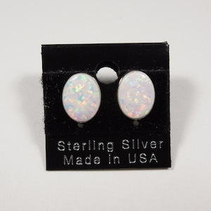May include: Oval-shaped opal stud earrings set in sterling silver. The opals display a mix of white, iridescent colors. The earrings are displayed on a black card that reads "Sterling Silver Made in USA".