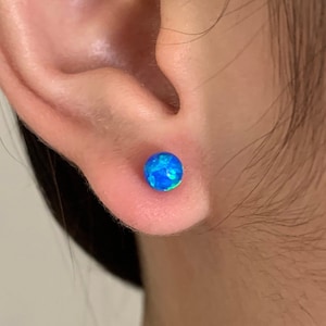 May include: A close-up of a person's ear with a blue opal stud earring. The earring is a round, flat disc with a blue and green iridescent finish.
