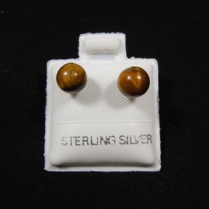 Pode incluir: Um par de brincos de prata esterlina com pedras de olho de tigre. Os brincos são exibidos em um cartão branco com as palavras "STERLING SILVER" impressas nele.