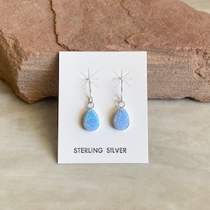 May include: A pair of sterling silver earrings with a teardrop-shaped, light blue opal stone. The earrings are displayed on a white card with the words "STERLING SILVER" printed in black. The earrings have a simple hook design.
