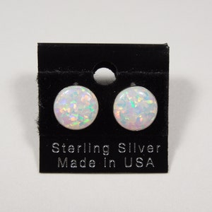 Puede incluir: Un par de pendientes de botón de ópalo blanco en una tarjeta negra con el texto "Sterling Silver Made in USA".