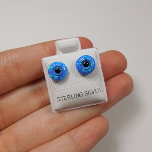 Puede incluir: Un par de pendientes de botón de ópalo azul con postes de plata de ley. Los pendientes están en una tarjeta blanca con las palabras "Sterling Silver" impresas en ella.