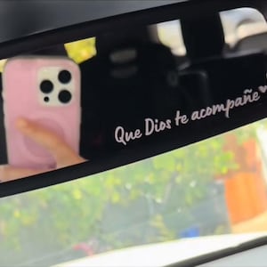 May include: A rearview mirror with a pink phone case and a sticker that reads "Que Dios te acompañe" with a small heart symbol.