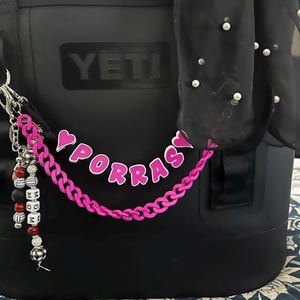 May include: Black cooler bag with a pink chain and the word "PORRAS" in pink letters. A beaded keychain with black, white, and red beads hangs from the bag. A black scarf with white pearls is draped over the bag.