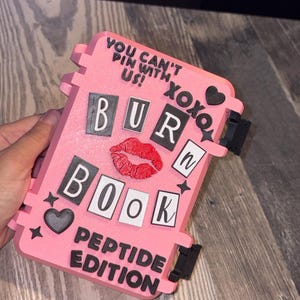 May include: A pink rectangular case with black and white lettering that reads YOU CAN'T PIN WITH US! XOXO, BURN BOOK, and PEPTIDE EDITION. The case features a red lip graphic, black heart icons, and small star shapes on the front surface.