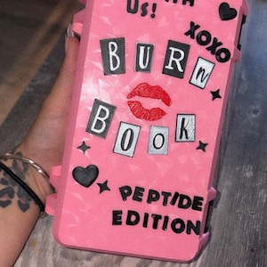 May include: A rectangular pink phone case featuring a textured geometric pattern. The text reads YOU CAN'T PIN WITH US! XOXO, BURN BOOK, and PEPTIDE EDITION. It includes a red lip print, black heart icons, and small black star accents.