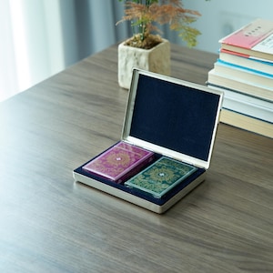 Puede incluir: Una caja de cartas de juego plateada con dos barajas de cartas dentro. Una baraja es verde con detalles dorados y la otra es rosa con detalles dorados.