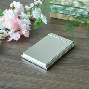 May include: A silver metal notebook with a blank page and a silver metal cover. The notebook is sitting on a wooden surface.