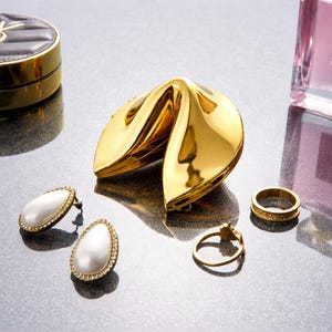 Op de afbeelding: Een goudkleurige sieradendoos in de vorm van een fortune cookie is het middelpunt, omringd door gouden ringen en parel oorbellen. Een zwart en goud compact poeder en een roze parfumfles zijn ook zichtbaar. De items zijn geplaatst op een reflecterend oppervlak.