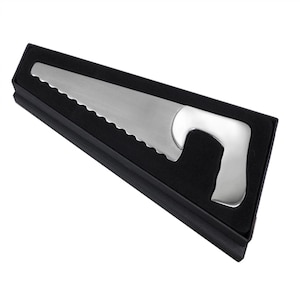 May include: A silver bottle opener in the shape of a saw, displayed in a black box. The opener features a serrated edge and a curved handle. The box is rectangular and has a black interior.