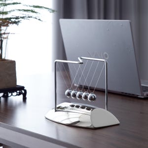Op de afbeelding: Een verchroomde Newton's cradle, een bureauspeelgoed met vijf zwaaiende metalen bollen. Het zilveren frame staat op een bijpassende basis. Op de achtergrond is een laptop te zien, wat wijst op een werkomgeving.