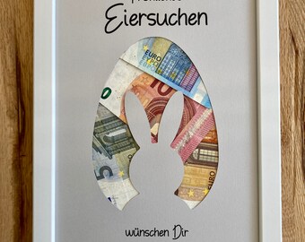 May include: A white picture frame with a gray background. The frame has a cutout of a bunny rabbit with euro bills inside. The text "Fröhliches Eiersuchen" is at the top of the frame and the text "wünschen Dir Tante Inge & Onkel Werner" is at the bottom.