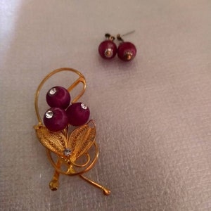 May include: A gold-tone brooch with three burgundy-colored beads and a filigree design. The brooch is accompanied by a pair of matching earrings with burgundy-colored beads.