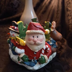 May include: A ceramic Christmas candle holder featuring Santa Claus, a snowman, and a reindeer. The figures are painted in bright colors and have a whimsical, festive look.