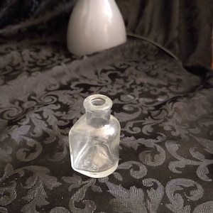 May include: A clear glass bottle with a square shape and a rounded top. The bottle is sitting on a black fabric with a floral pattern.