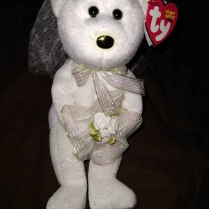 May include: White plush bear wearing a white veil and holding a bouquet of white roses. The bear has a gold nose and black eyes. The bear is wearing a pink ribbon around its neck. The bear has a Ty Beanie Babies tag attached to its ear.