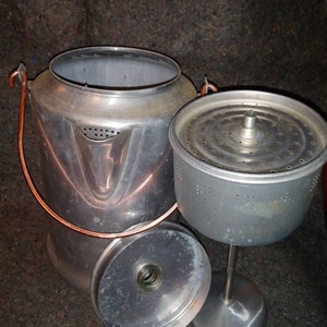 May include: A vintage silver-colored percolator coffee pot with a copper handle and a removable filter. The pot has a round base and a tall, cylindrical body.