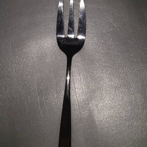 May include: A silver fork with four tines. The fork is on a gray surface.