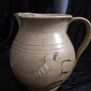 May include: A brown stoneware pitcher with a wide mouth and a handle. The pitcher has a speckled glaze and a brown and white design.