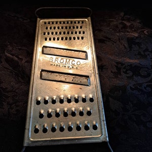 May include: A silver metal grater with a black handle. The grater has a rectangular shape and is labeled "BROMCO MADE IN U.S.A."
