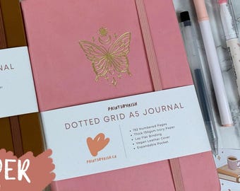 May include: A pink A5 dotted grid journal with a gold butterfly design and the text "PRINTSBYRISH DOTTED GRID A5 JOURNAL" on the front cover. The journal has 192 numbered pages, thick 150gsm ivory paper, lay flat binding, vegan leather cover, and an expandable pocket.