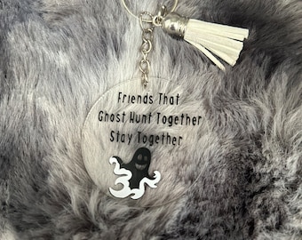 Może przedstawiać: Brelok do kluczy z białym frędzlem, srebrnym łańcuszkiem i przezroczystym okrągłym breloczkiem z napisem "Friends That Ghost Hunt Together Stay Together" i czarnym rysunkiem ducha.