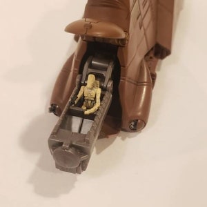 May include: A brown plastic toy vehicle with a figure of a Star Wars character sitting inside. The vehicle has a raised canopy and a long, cylindrical gun.