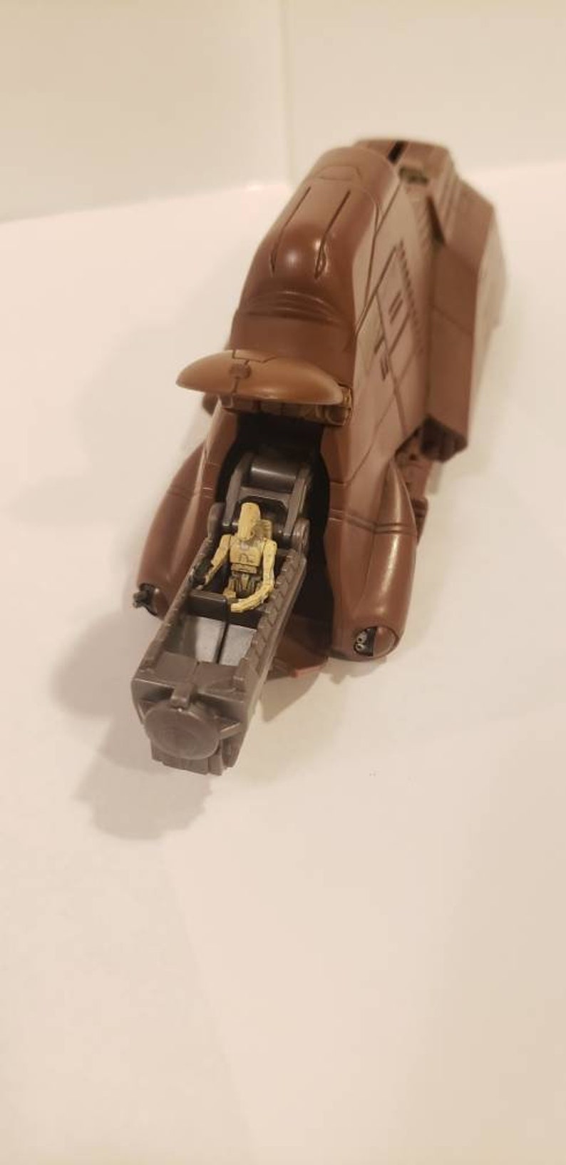 May include: A brown plastic toy vehicle with a figure of a Star Wars character sitting inside. The vehicle has a raised canopy and a long, cylindrical gun.