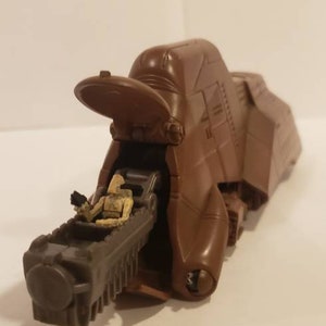 May include: A brown plastic Star Wars vehicle with a figure of a sandtrooper sitting inside. The vehicle has a large, open cockpit and a long, articulated arm.