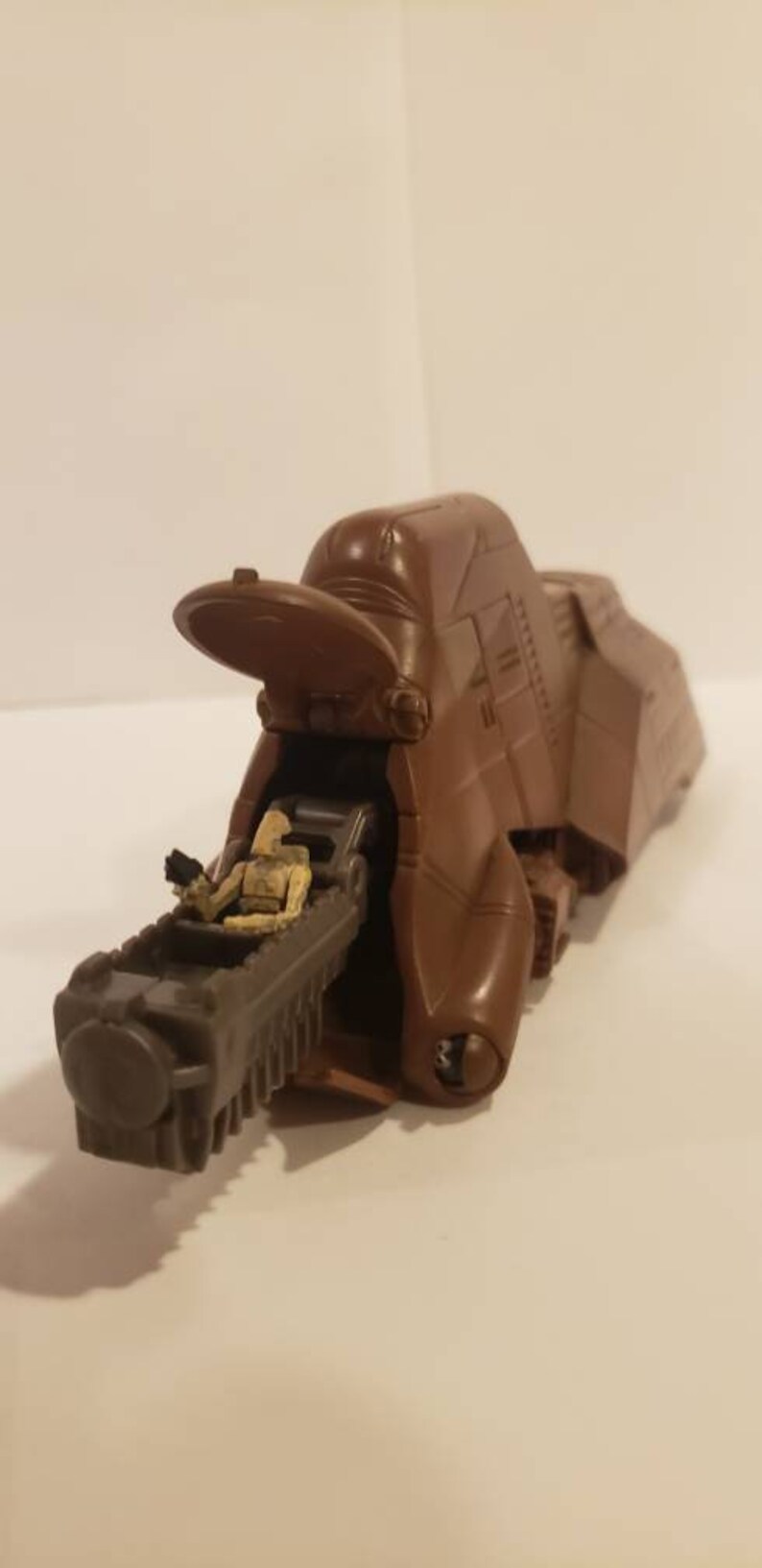 May include: A brown plastic Star Wars vehicle with a figure of a sandtrooper sitting inside. The vehicle has a large, open cockpit and a long, articulated arm.