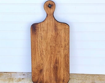 May include: A rectangular wooden cutting board with a handle. The board is brown and has a natural wood grain. The handle is round and has a hole in the top.