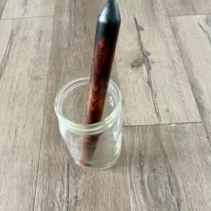 May include: A clear glass jar with a dark wooden rolling pin inside. The rolling pin has a black tip and a reddish-brown body. The jar is sitting on a light brown wooden surface.